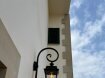 Wandleuchte aussen von Roger Pradier. Der Wandarm ist gebogen, das Lampengehäuse viereckig, unten geöffnet. Die Lampe hat vier Lampengläser, Das Oberteil ist geschlossen. Montiert an Bischofsstab an mediterranem Wandhaus.