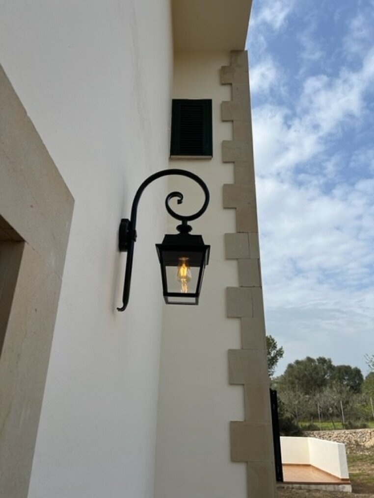 Wandleuchte aussen von Roger Pradier. Der Wandarm ist gebogen, das Lampengehäuse viereckig, unten geöffnet. Die Lampe hat vier Lampengläser, Das Oberteil ist geschlossen. Montiert an Bischofsstab an mediterranem Wandhaus.