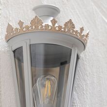 Die Wandlaterne Louvre Modell 4 zeigt ihre Detailstärke in dieser Nahaufnahme. Der graue Metallrahmen wird durch eine kunstvoll gestaltete goldene Krone ergänzt, die der Leuchte eine elegante und luxuriöse Note verleiht. Klare Glaseinsätze umrahmen die moderne LED-Birne, die für eine gleichmäßige Lichtverteilung sorgt. Diese Leuchte ist ideal für dekorative Akzente an Fassaden und Eingangsbereichen und besticht durch ihre hochwertige Verarbeitung.