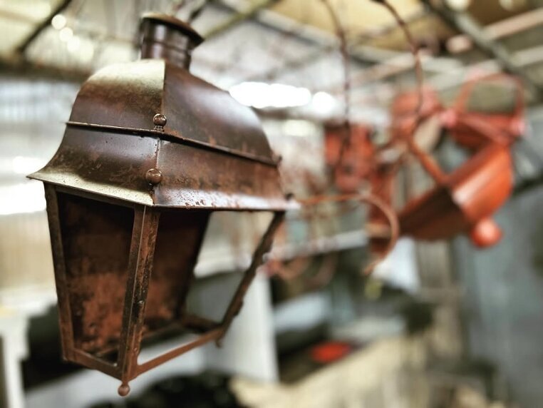 Die Mazarine Wall Lantern zeigt sich hier im Rostfinish während des handwerklichen Entstehungsprozesses. Die Oberfläche ist noch nicht vollendet und offenbart bereits die charakteristische Tiefe aus warmen Braun- und Kupfertönen, die durch kontrollierte Oxidation entstehen. Feine Unebenheiten und natürliche Farbverläufe unterstreichen den authentischen Manufakturcharakter der klassischen Wandlaterne. Im Hintergrund sind weitere Leuchten im Arbeitsprozess zu erkennen, was die sorgfältige, manuelle Fertigung und die traditionsreiche Verarbeitung dieser französisch inspirierten Außenleuchte eindrucksvoll dokumentiert.