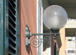 Wandleuchte mit Glaskugel am Jugendstil Wandarm. anthrazit-grau. An einem italienischen Stadthaus mit Terrakotta Mauerwerk und grünen Fensterläden. Lampenglas als satinierte Kugel.