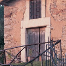 Die mediterrane Wandleuchte Napoli Nr. 21110 heißt Gäste mit warmem Licht willkommen. Runde Glaskugel an gebogenem Wandarm vor einem Denkmal geschützen Gebäude