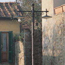 schöne mediterrane Gartenleuchte mit 2 lampenauslegern. Schmaler Mast, Wandarme Jugendstil. Die Aussenleuchte steht vor typisch italienischer haeuser kulisse. Natursteinmauern.