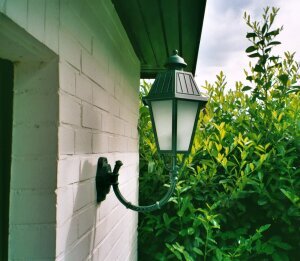 Wandleuchte mit sechseckigem Wandarm, der gebogen ist. Befestigt an weißem Klinkerhaus.