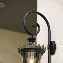 Die gezeigte Wandlaterne vereint klassische Eleganz mit hochwertigem Design. Der schwarze Metallrahmen wird durch eine kunstvoll gestaltete goldene Krone veredelt. Die klare Glaseinfassung zeigt die Glühbirne im Inneren und sorgt für eine harmonische Lichtverteilung. Die dekorative geschwungene Wandhalterung ergänzt das Design perfekt. Ideal für historische Fassaden oder stilvolle Außenbereiche.