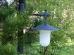 Der Lampenmast ist rund mit einem runde Befstigungsfuß. Der Arm ist im Jugendstil gehalten, die Lampe ist rund und oben in der Farbe grau, unten weiß. Das Lampenglas ist rund.