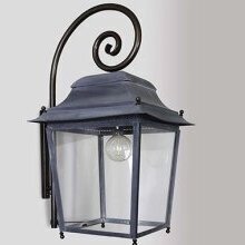 Historische Außenwandleuchte Chantilly Nr. 1018 Géante im aged zinc-Finish an einer weißen Putzfassade. Man sieht den viereckigen Lampenkörper aus gealgtertem Zink an einem gebogenen Wandarm in Form eines Bischofsstab.