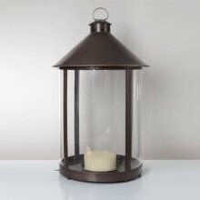 Seitliche Detailaufnahme der handgefertigten Außenlampe Luxembourg mit geschwungener Silhouette und Glaszylinder. Die Leuchte hat ein rundes Design und steht auf dem Boden vor einer Hauswand.