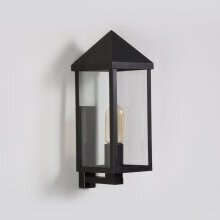 Starker Kontrast – geometrisches Lichtobjekt in natürlicher Umgebung. Kurzer Wandarm mit rechteckigem Gehäuse und spitzem Dach, schwarze Farbe mit Klarglasscheiben.