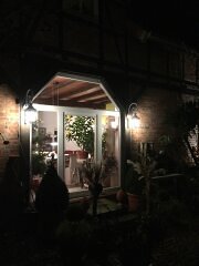 weiße, sechseckige Wandlampen mit klarem Glas am Bischofsstab.Rechts und links von großem, weißen Deelen Fenster. Es ist dunkel und dieAußenleuchten sind eingeschaltet.