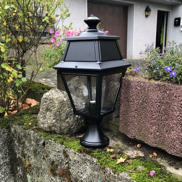 schwarze kleine Wegeleuchte mit Klarglas steht in einem Beet mit braunem Laub. Dahinter sind grüne Blätter von Sträuchern.